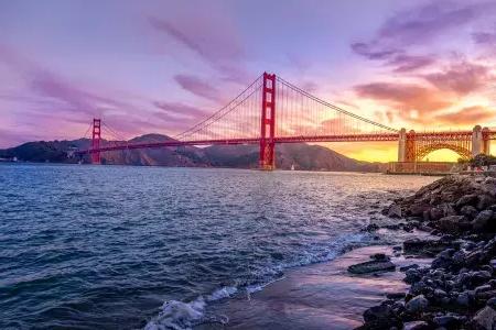 The golden gate bridge at sunset with a multicolored sky and the San Francisco Bay in the foreground.